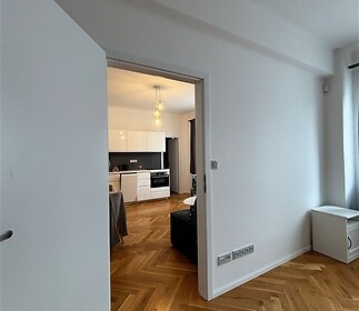 Vstup do moderní kuchyně s parketovou podlahou, bílou kuchyňskou linkou a velkými okny, které propouštějí spoustu světla.