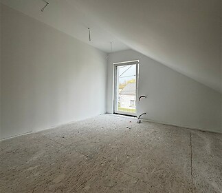 Podkrovní pokoj s OSB podlahou, připravený k dokončení. Světlé stěny, šikmá střecha a velké okno dodávají místnosti moderní vzhled.