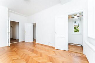 Světlý obývací pokoj s parketovou podlahou a bílými stěnami, moderní interiér, velká okna poskytující dostatek přirozeného světla.