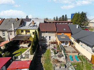 Zahrada s rekonstruovaným domem, dřevěnou terasou a malým trávníkem, obklopená živými ploty a stromy, nabízí klidné prostředí pro odpočinek.