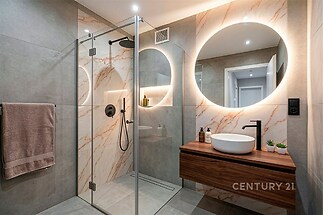 Moderní koupelna s elegantními dlaždicemi, sprchovým koutem a osvětleným zrcadlem, doplněná o moderní umyvadlo na dřevěné desce.