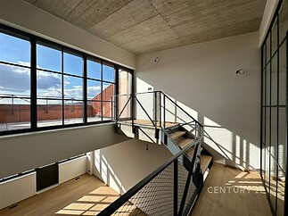 Světlé schodiště s moderním kovovým zábradlím, velká okna a parketová podlaha umožňují přísun přirozeného světla v industriálním stylu.