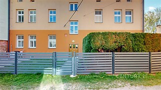 Fasáda bytového domu s moderním oplocením, velkými okny a udržovanou zahradou, ideální pro klidné bydlení ve městě.