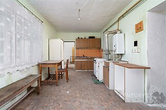 Kuchyně s dřevěným nábytkem, dlažbou a velkými okny, vyžadující rekonstrukci, nabízí potenciál pro modernizaci a zlepšení.