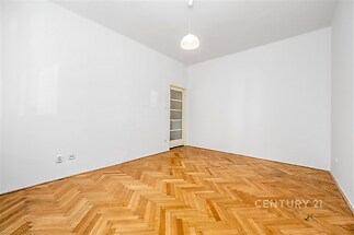 Prázdný pokoj s parketovou podlahou, bílými stěnami a moderním osvětlením. Vstupní dveře s prosklením zajišťují světlo do prostoru.