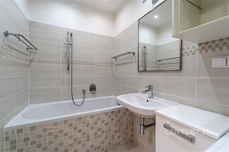 Moderní koupelna s dlažbou, světlými obklady a vestavěnou vanou. Elegantní zrcadlo a praktické úložné prostory.