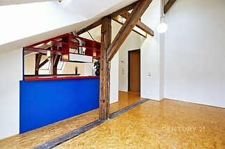 Prostorný obývací pokoj s parketovou podlahou, odkrytými dřevěnými trámy a moderní vestavěnou kuchyňskou linkou v modré a červené barvě.