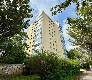 Vysoký bytový dům s prosklenými balkony, obklopený zelení a umístěný v klidné městské lokalitě.