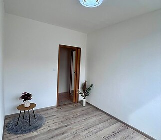 Prázdná místnost s dřevěnou podlahou a bílými stěnami, moderní svítidlo, výrazné světlo, dveře vedoucí na dlažbu v chodbě.