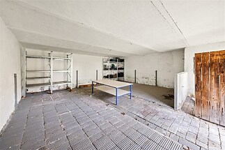Garáž s betonovou podlahou, vybavená regály a pracovní lavicí, vhodná pro rekonstrukci. Prostorný interiér nabízí možnosti úložného prostoru.