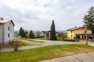 Výhled z balkonu na klidnou rezidenční čtvrť s rodinnými domy, udržovanou zahradou a dlážděnými chodníky, vhodné pro rodiny hledající klidné bydlení.