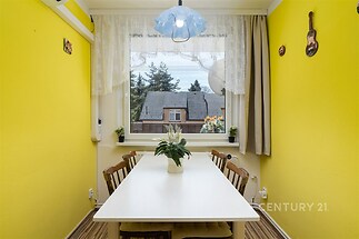 Jídelna s dřevěnou podlahou a velkými okny, světlý interiér s moderním nábytkem, dekorativní prvky na stěnách přidávají útulnost.