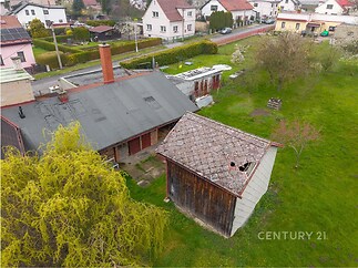 Starší zahradní kůlna s poškozenou střešní krytinou se nachází na rozlehlém pozemku, kolem jsou vzrostlé stromy a přilehlé domy.