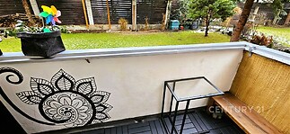 Malý balkon s dřevěnou podlahou, zdobený mandalou na stěně, s výhledem na zahradu. Obsahuje minimalistický nábytek a květinové dekorace.