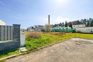Prázdná stavební parcela s travnatým povrchem, poblíž průmyslových budov a komína, ideální pro výstavbu nebo investici, slunná oblast.