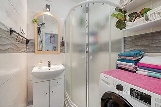 Malá koupelna s moderním sprchovým koutem, světlé dlažby, vestavěné police, pračka, čistý a udržovaný prostor.