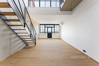 Světlý obývací pokoj s parketovou podlahou, velká okna a moderní schodiště. Industriální styl a vysoké stropy vytvářejí prostorný dojem.