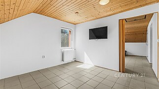 Světlý pokoj s dlažbou, dřevěným stropem a velkými okny. Moderní úpravy a čisté linie vytvářejí prostorný a světlý interiér.