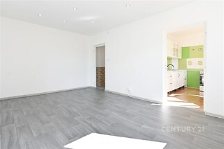 Světlý obývací pokoj s parketovou podlahou, moderní osvětlení a vstup do kuchyně s dlažbou, renovovaný prostor vhodný pro rodinné bydlení.