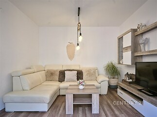 Světlý obývací pokoj s dřevěnou podlahou, moderním nábytkem a velkým rohovým gaučem, dekorativní osvětlení dodává útulný pocit.