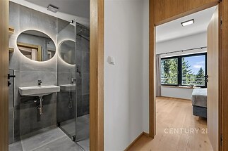 Chodba s dřevěnými rámy vedoucí do moderní koupelny s dlažbou a sprchovým koutem a do světlé ložnice s parketovou podlahou a velkými okny.