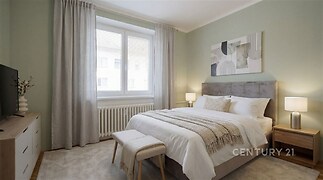 Světlá ložnice s velkým oknem, moderní nábytkem a neutrálními barvami, útulný koberec a minimalistický dekor vytvářejí příjemnou atmosféru.