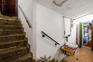 Spodní část schodiště s kamennými schody, omítkovými stěnami a dlažbou na podlaze, v chodbě je také umístěna pračka.