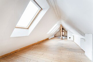 Podkrovní místnost s dřevěnou podlahou, velkými střešními okny a bílými stěnami, nabízí světlý prostor vhodný k renovaci.