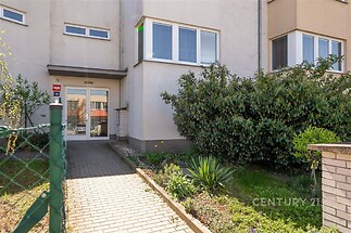 Vchod do bytového domu s upravenou zahrádkou, zajišťující soukromí a bezpečí. Cihlová dlažba a moderní vstupní dveře.