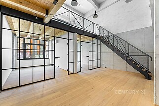 Prostorný moderní obývací pokoj s parketovou podlahou, velkými prosklenými stěnami a kovovým schodištěm, ideální pro loftový styl.