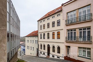 Historická fasáda bytového domu na klidné ulici s dlážděným povrchem, nabízí renovovaný prostor s velkými okny pro příjemné bydlení.