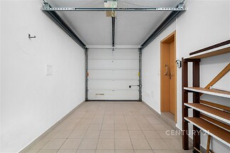 Garáž s dlažbou a bílými stěnami, vybavená kovovými regály a automatickými vraty, ideální pro skladovací využití nebo parkování