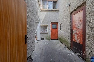 Úzký venkovní dvorek se vstupními dveřmi s dřevěnou texturou, omítnutými stěnami a zastřešením, ideální pro skladování nebo renovaci.