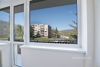 Světlý balkonový prostor s velkými okny a moderním vybavením, nabízí výhled na udržovanou zahradu a okolní budovy, ideální pro relaxaci