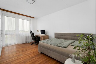 Světlá ložnice s laminátovou podlahou, velká okna s bílými závěsy, moderní nábytek a pracovní stůl, ideální pro klidné bydlení.