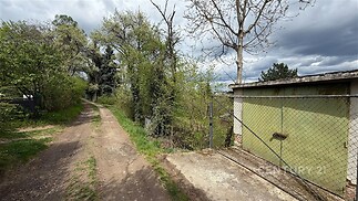 Příjezdová cesta s nezpevněným povrchem vedle zahrady. K pozemku patří garáž s plechovými dveřmi, obklopená zelení a stromy.