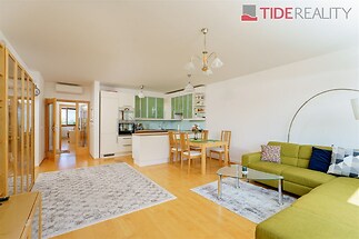 Světlý obývací pokoj s parketovou podlahou, otevřený do moderní kuchyně, vybavený velkými okny a komfortní sedací soupravou.