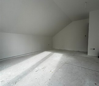 Nezařízená podkrovní místnost s vysokým stropem, betonová podlaha, bíle vymalované stěny, světlý prostor připravený k dokončení