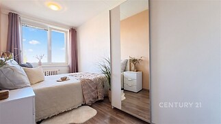 Světlá ložnice s parketovou podlahou, velkým oknem a moderním nábytkem včetně vestavěné skříně, nabízí pohodlný obytný prostor.