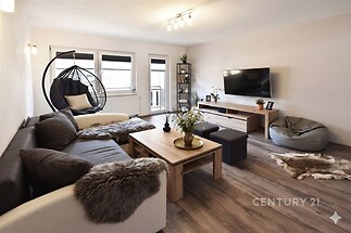 Světlý obývací pokoj s moderním designem, dřevěnou podlahou a velkými okny, vybavený pohodlnou sedací soupravou a nástěnnou televizí.