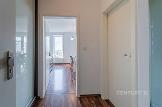 Světlá chodba s parketovou podlahou vede do moderního obývacího pokoje s velkými okny a bílými zdmi.