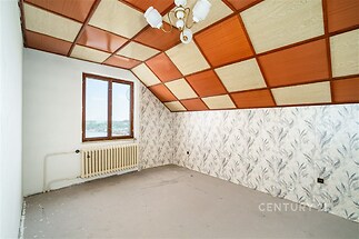 Podkrovní místnost s dřevěným obložením, velkým oknem a tapetami, vyžadující rekonstrukci, nabízí potenciál pro světlý interiér.
