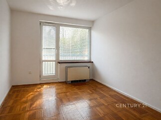 Světlý pokoj s parketovou podlahou, velkým oknem a moderním vytápěcím tělesem, ideální pro minimalistický styl.