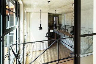 Dvoupatrový loft s dřevěnou podlahou, velkými okny a moderními svítidly nabízí světlý a prostorný interiér s industriálním nádechem.