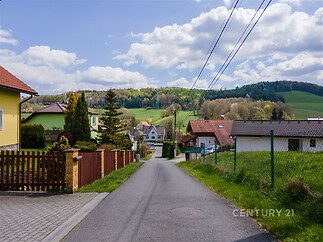 Pohled na klidnou rezidenční ulici v malebné oblasti s rodinnými domy obklopenými zelení a kopci v pozadí.