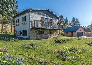 Útulný venkovský dům s kamennou fasádou, dřevěný balkon s květinami, rozkvetlá zahrada a velká okna pro světlý interiér.