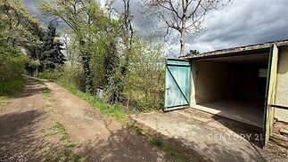 Prázdná garáž s betonovou podlahou a dřevěnými vraty, venku nezpevněná cesta obklopená zelení, vhodné pro skladování nebo parkování.