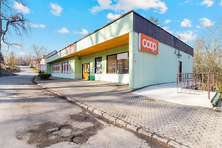 Exteriér komerční budovy s moderní fasádou, velkými okny a vstupními dveřmi. Budova je obklopena zpevněnou plochou a přístupovou cestou.