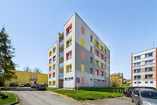 Bytový dům se světlou fasádou a barevnými detaily, moderní architektura, klidná lokalita, venkovní parkování, blízkost zeleně.