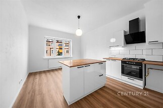 Moderní kuchyň s parketovou podlahou, světlá díky velkým oknům, vybavená vestavěnými spotřebiči a elegantními bílými skříněmi.